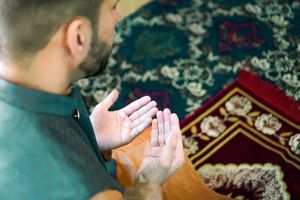 A man in traditional attire is praying on a beautifully decorated prayer mat indoors.