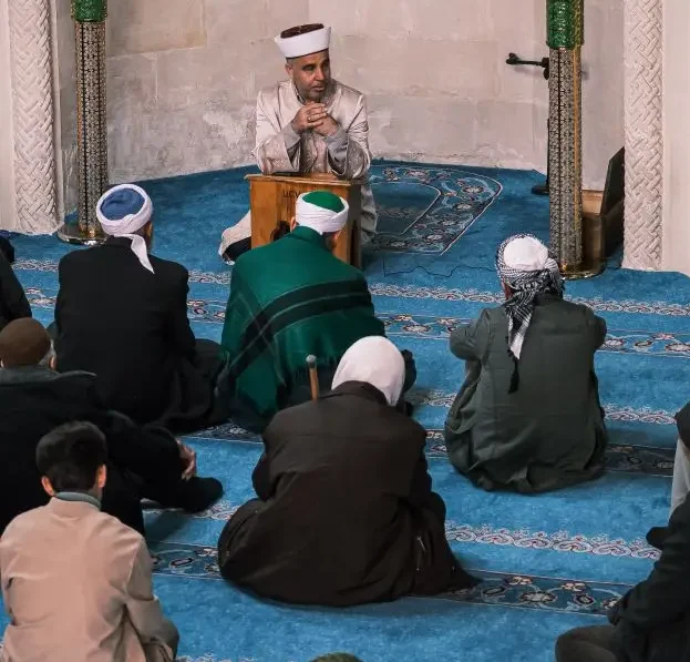 An imam leads a group prayer session in a mosque, surrounded by worshippers on blue carpeted floor.