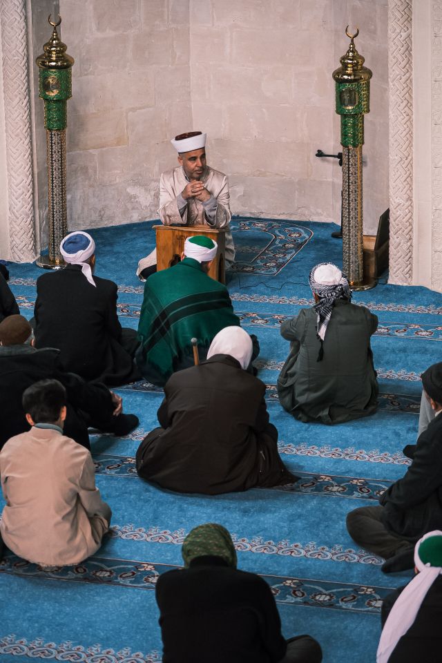 An imam leads a group prayer session in a mosque, surrounded by worshippers on blue carpeted floor.